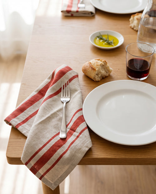 A Pomodoro Righe napkin beside a white plate at a Sunday lunch table, with bread, olive oil and red wine